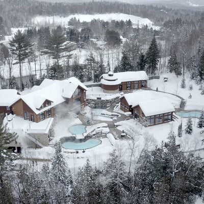 Vue aérienne du complexe complet de l'Amerispa Spa Nordique Morin-Heights en hiver, niché dans la forêt laurentienne.