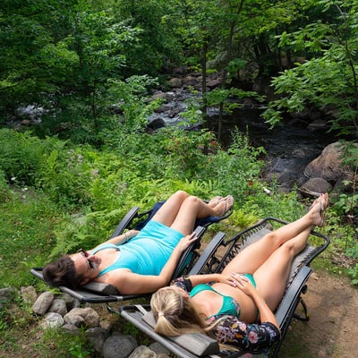 Deux femmes se relaxant sur des chaises longues directement au bord de la rivière aux Mulets, dans l'enceinte de l'Auberge & Spa Beaux Rêves.