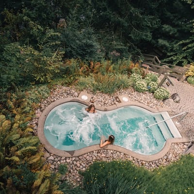 Vue aérienne de deux personnes se relaxant dans un bain chaud au Scandinave Spa Mont-Tremblant, entouré de fougères et de la forêt laurentienne.