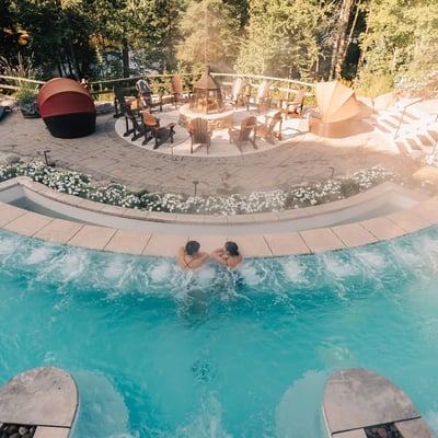 Vue de dessus d'un couple se relaxant dans une piscine au Scandinave Spa, avec l'aire de détente et le foyer extérieur en arrière-plan.