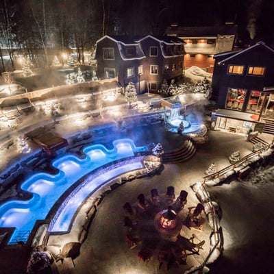 Vue aérienne nocturne du Scandinave Spa en hiver, avec les bains illuminés en bleu, le foyer allumé et le site couvert de neige.