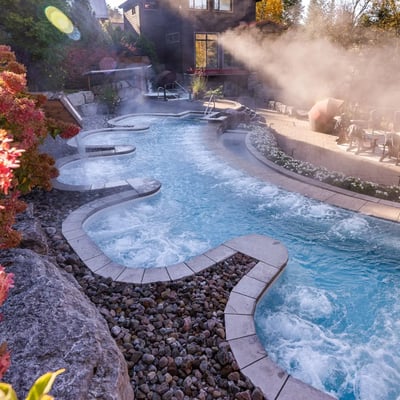 Les bains à remous du Scandinave Spa dégageant de la vapeur au petit matin, bordés de fleurs et de roches naturelles.