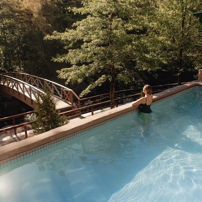 Femme se relaxant dans un bain extérieur au Strøm Spa de Saint-Sauveur, avec une vue sur le pont en bois qui enjambe la rivière.