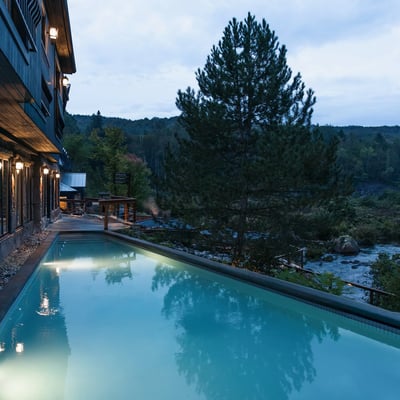 Piscine extérieure illuminée du Strøm Spa de Saint-Sauveur au crépuscule, surplombant la rivière et la forêt.