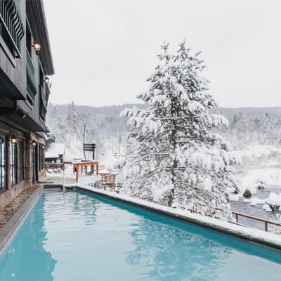 Piscine extérieure chauffée du Strøm Spa Nordique Saint-Sauveur en hiver, avec vue sur la rivière et la forêt enneigées.