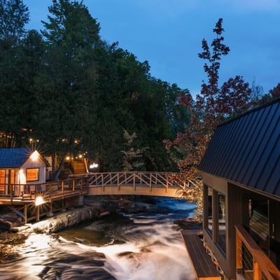 Vue en soirée du Strøm Spa Nordique à Saint-Sauveur, avec la rivière à Simon illuminée et le pont en bois reliant les installations.